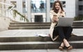 Young beautiful businesswoman having phone conversation works in her laptop drinks coffee sitting on stairs near office center in Royalty Free Stock Photo