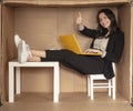 Young beautiful businesswoman sitting in office cardboard box, happy shows thumbs up