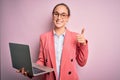 Young beautiful businesswoman wearing glasses working using laptop over pink background happy with big smile doing ok sign, thumb Royalty Free Stock Photo