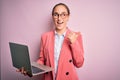 Young beautiful businesswoman wearing glasses working using laptop over pink background pointing and showing with thumb up to the Royalty Free Stock Photo