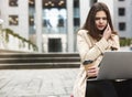 Young beautiful businesswoman works in her laptop drinks coffee sitting on stairs near office center in downtown, looks upset, Royalty Free Stock Photo