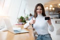 Young beautiful businesswoman works in her laptop and holds bank card showing like sign while sitting in the cafe during lunch Royalty Free Stock Photo