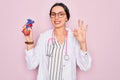 Young beautiful cardiologist woman with blue eyes wearing stethoscope holding heart doing ok sign with fingers, excellent symbol