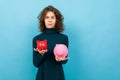 Young and beautiful caucasian girl with curly hair keeps small red pig moneybox, big pink pig moneybox and smiles