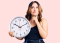 Young beautiful caucasian woman holding big clock serious face thinking about question with hand on chin, thoughtful about Royalty Free Stock Photo