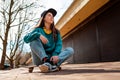 A young beautiful Caucasian woman sits relaxed on a skateboard and looks away. In the background, a street with trees. Bottom view