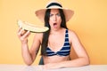 Young beautiful caucasian woman wearing bikini and hat holding slice of melon scared and amazed with open mouth for surprise, Royalty Free Stock Photo