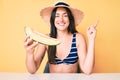 Young beautiful caucasian woman wearing bikini and hat holding slice of melon smiling happy pointing with hand and finger to the Royalty Free Stock Photo