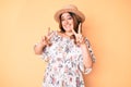 Young beautiful caucasian woman wearing summer dress and hat smiling looking to the camera showing fingers doing victory sign Royalty Free Stock Photo