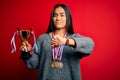 Young beautiful champion asian woman holding trophy wearing medals over red background with angry face, negative sign showing Royalty Free Stock Photo