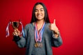 Young beautiful champion asian woman holding trophy wearing medals over red background happy with big smile doing ok sign, thumb Royalty Free Stock Photo