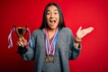 Young beautiful champion asian woman holding trophy wearing medals over red background very happy and excited, winner expression