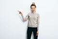 Young beautiful cheerful woman with hair bun wearing in jeans and sweater pointing with forefinger on copy space over white wall Royalty Free Stock Photo