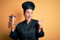 Young beautiful chef woman wearing cooker uniform and hat holding bottle with italian pasta pointing and showing with thumb up to Royalty Free Stock Photo