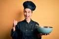 Young beautiful chef woman wearing cooker uniform and hat holding bowl and whisk pointing and showing with thumb up to the side Royalty Free Stock Photo