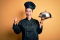 Young beautiful chef woman wearing cooker uniform and hat holding tray with dome pointing and showing with thumb up to the side Royalty Free Stock Photo