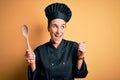 Young beautiful chef woman wearing cooker uniform and hat holding wooden spoon pointing and showing with thumb up to the side with Royalty Free Stock Photo