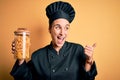 Young beautiful chef woman wearing cooker uniform holding jar with italian pasta macaroni pointing and showing with thumb up to Royalty Free Stock Photo