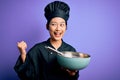 Young beautiful chinese chef woman wearing cooker uniform and hat using bowl and whisk pointing and showing with thumb up to the Royalty Free Stock Photo