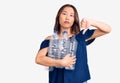 Young beautiful chinese girl holding recycling plastic bottles with angry face, negative sign showing dislike with thumbs down,