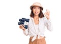 Young beautiful chinese girl wearing explorer hat holding binoculars doing ok sign with fingers, smiling friendly gesturing Royalty Free Stock Photo