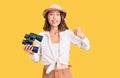 Young beautiful chinese girl wearing explorer hat holding binoculars smiling happy and positive, thumb up doing excellent and Royalty Free Stock Photo