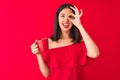 Young beautiful chinese woman drinking cup of coffee standing over isolated red background with happy face smiling doing ok sign Royalty Free Stock Photo