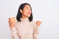 Young beautiful chinese woman drinking paper glass of coffee over isolated white background pointing and showing with thumb up to Royalty Free Stock Photo