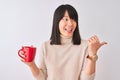 Young beautiful Chinese woman drinking red cup of coffee over isolated white background pointing and showing with thumb up to the Royalty Free Stock Photo