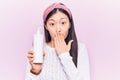 Young beautiful chinese woman holding bottle with cream lotion covering mouth with hand, shocked and afraid for mistake