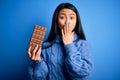 Young beautiful chinese woman holding chocolate bar over isolated blue background cover mouth with hand shocked with shame for Royalty Free Stock Photo