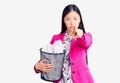 Young beautiful chinese woman holding paper bin full of crumpled papers pointing with finger to the camera and to you, confident