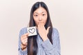 Young beautiful chinese woman holding reminder with heart shape covering mouth with hand, shocked and afraid for mistake
