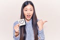 Young beautiful chinese woman holding reminder with heart shape pointing thumb up to the side smiling happy with open mouth