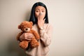 Young beautiful chinese woman holding teddy bear standing over isolated white background cover mouth with hand shocked with shame Royalty Free Stock Photo