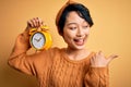 Young beautiful chinese woman holding vintage alarm clock over isolated yellow background pointing and showing with thumb up to Royalty Free Stock Photo
