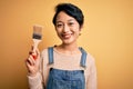 Young beautiful chinese woman painting holding paint brush over isolated yellow background with a happy face standing and smiling Royalty Free Stock Photo
