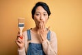 Young beautiful chinese woman painting holding paint brush over  yellow background cover mouth with hand shocked with Royalty Free Stock Photo