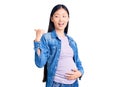 Young beautiful chinese woman pregnant expecting baby smiling with happy face looking and pointing to the side with thumb up