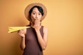 Young beautiful chinese woman on vacation wearing summer hat holding paper airplane cover mouth with hand shocked with shame for