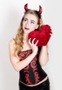 Young and beautiful curly girl with red horns looks like pretty Devil, holding a heart pillow Royalty Free Stock Photo