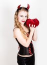 Young and beautiful curly girl with red horns looks like pretty Devil, holding a heart pillow Royalty Free Stock Photo