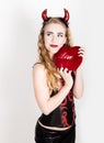 Young and beautiful curly girl with red horns looks like pretty Devil, holding a heart pillow Royalty Free Stock Photo