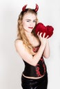 Young and beautiful curly girl with red horns looks like pretty Devil, holding a heart pillow Royalty Free Stock Photo