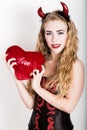 Young and beautiful curly girl with red horns looks like pretty Devil, holding a heart pillow Royalty Free Stock Photo