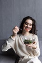 Young and beautiful woman sitting on sofa at home, eating colorful fresh vegetable salad and smiling, healthy woman enjoying food Royalty Free Stock Photo