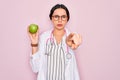 Young beautiful doctor woman with blue eyes wearing stethoscope holding green apple fruit pointing with finger to the camera and Royalty Free Stock Photo