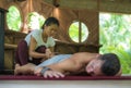 Young beautiful and exotic Asian Indonesian therapist woman giving traditional Thai massage to man relaxed at tropical wellness