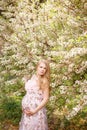 Young beautiful expectant mother hugs her hands belly near flowering trees