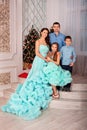 A young beautiful family of four in elegant dresses with a dog are standing at home posing near the New Year tree